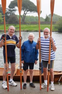 3 men stood on the bank beside a wooden row boat each holding two oars upright