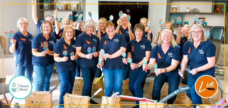 17 of the Sparkly Singers stood in a room smiling and holding paper maracas