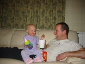 Isla as a toddler with a sippy cup as a baby sat on her father, who is led on the sofa holding a cup
