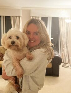 Isla in a living room, looking at the camera and holding a small white dog