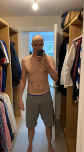 Iain stood in a walk in wardrobe. Wearing grey shorts with his shirt off taking a selfie in the mirror