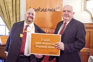 Joseph in a suit and red tie with MP Martin Rhodes both holding a plaque that says 'I will take action to improve lives'