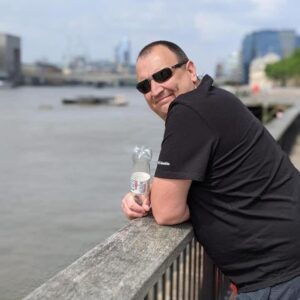 Martin Anderton by a river bank on a sunny day, leaning on a barrier and smiling at the camera
