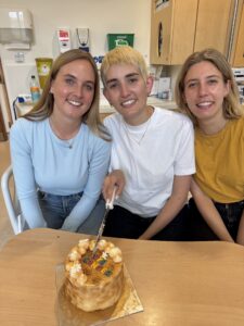 Alice holding a knife to a cake that says 'bye cancer' with her 2 sisters either side