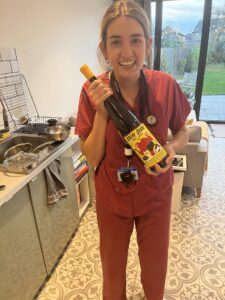 Alice stood in a kitchen in red hospital scrubs holding a bottle of wine called 'chin chin'